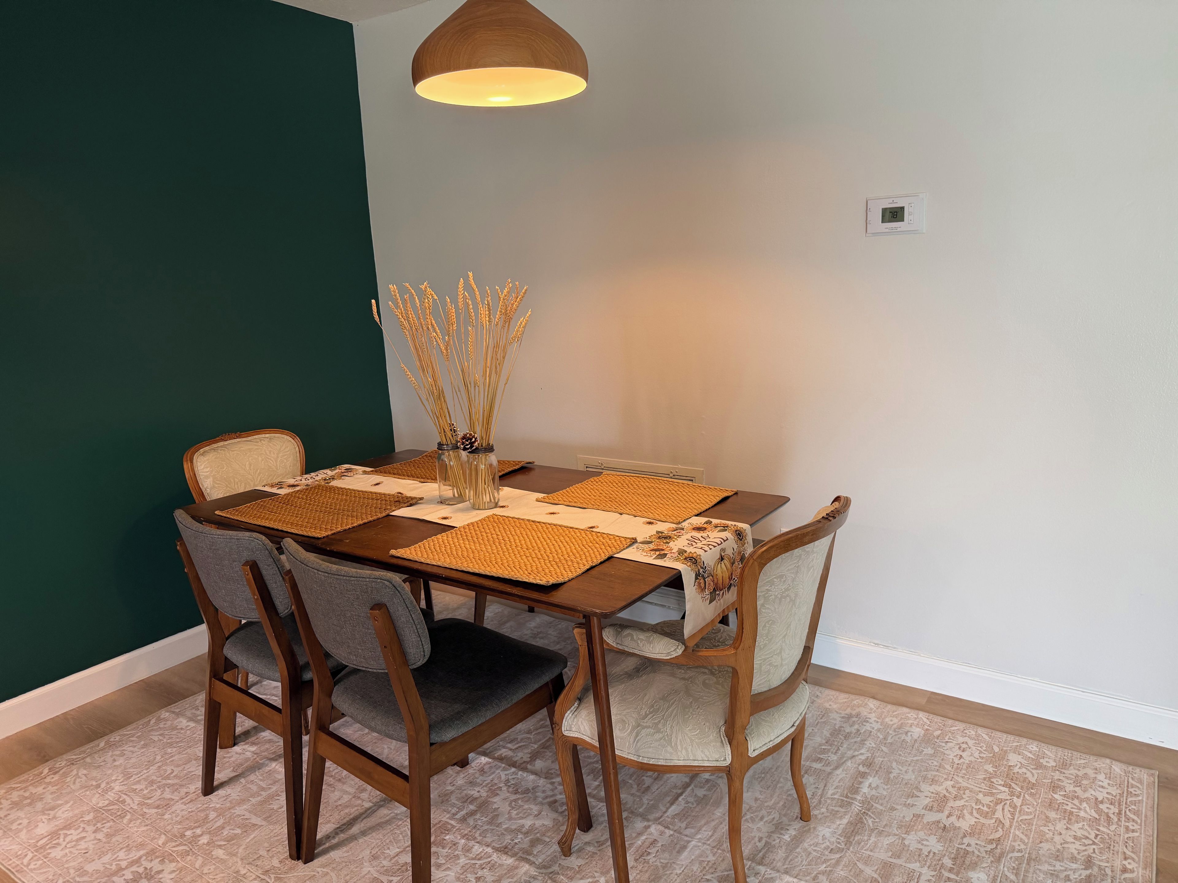 A dining area with a wooden table, two chairs, a vase with dried plants, and a pendant light.