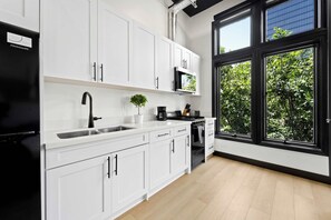 Eine moderne Küche mit weißen Schränken, einem schwarzen Kühlschrank, einer Kaffeemaschine und Blick ins Grüne durch große Fenster.
