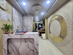 A reception area with a marble-patterned wall, a clock, and a ceiling fan. - Oly Hotel Yes Sir (New Delhi)