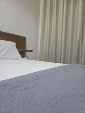 A bed with a white sheet and a grey blanket, a wooden headboard, and a curtain in the background. - SEED-Aeroport Nsimalen Yaounde (NSIMALEN)
