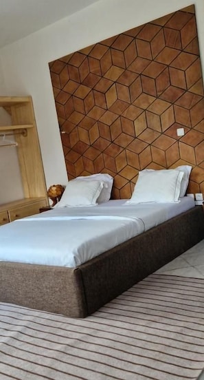 A bed with a headboard featuring a geometric pattern, a bedside table, and a striped rug. - SEED-Aeroport Nsimalen Yaounde (NSIMALEN)