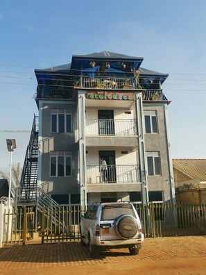 A multi-story building with balconies, a car parked in front, and a gated entrance.