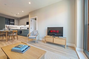 Ein modernes Wohnzimmer mit einem Fernseher, der 'NETFLIX' anzeigt, ein Essbereich mit Tisch und Stühlen sowie eine Küche im Hintergrund.