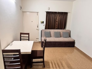 Una habitación con una mesa de comedor, sillas, un sofá con cojines estampados, una puerta y una ventana con cortinas.