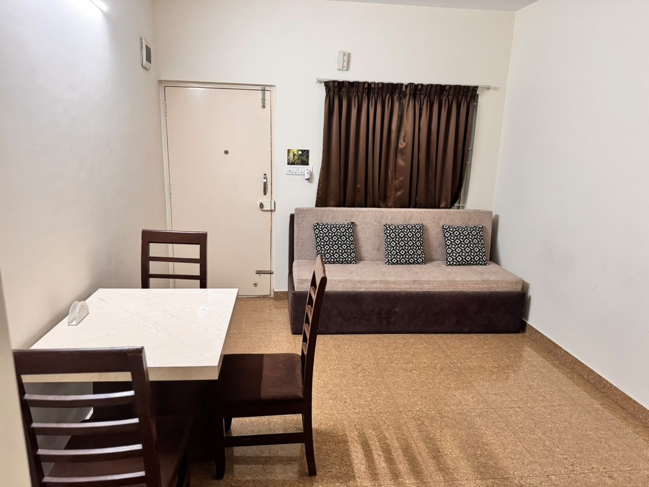 Una habitación con una mesa de comedor, sillas, un sofá con cojines estampados, una puerta y una ventana con cortinas.