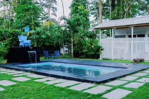 En swimmingpool med et vandfald, omgivet af frodig beplantning og et hvidt hegn.
