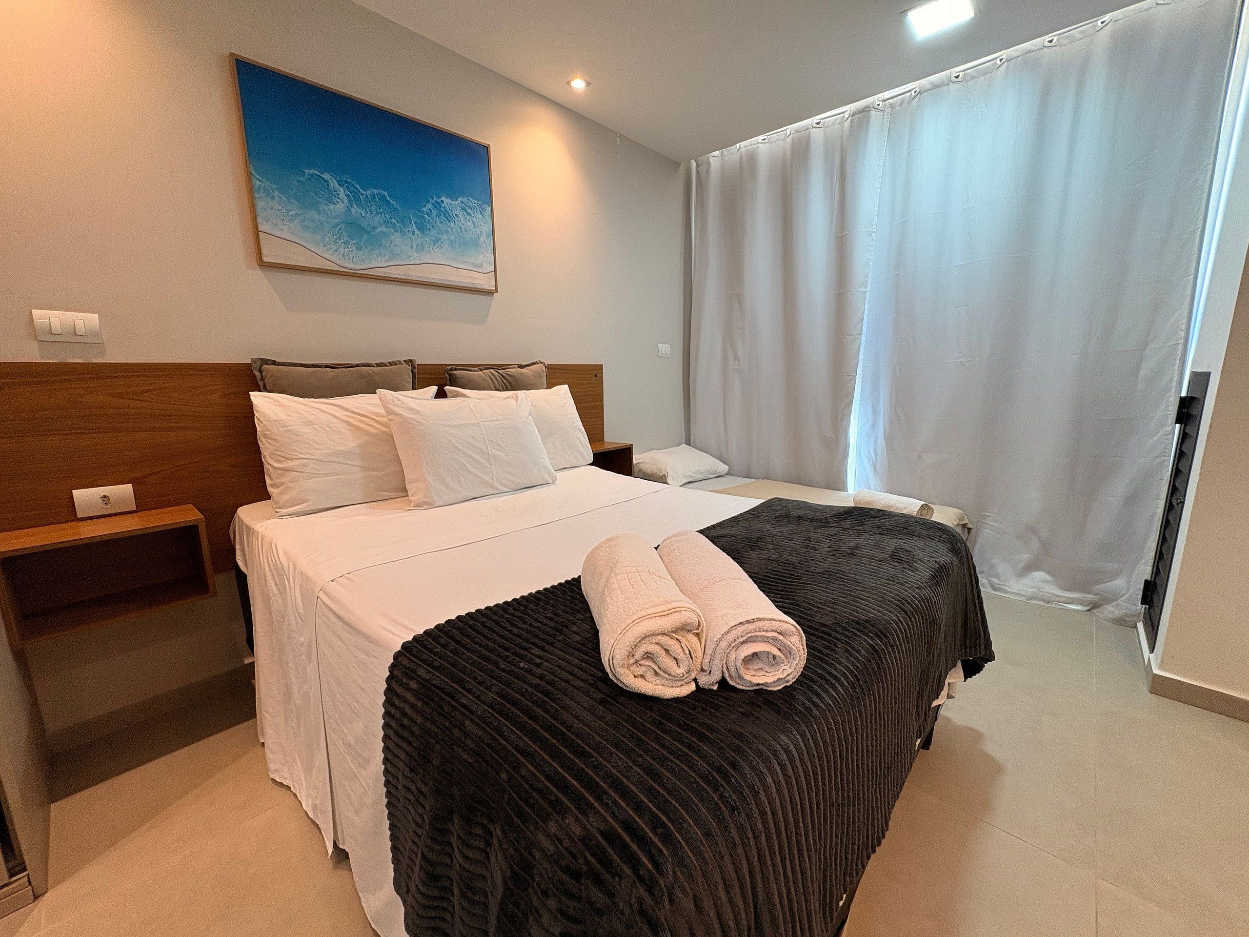 A hotel room with a bed, a nightstand, a painting, and a window with curtains.