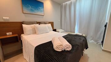 A hotel room with a bed, a nightstand, a painting, and a window with curtains.