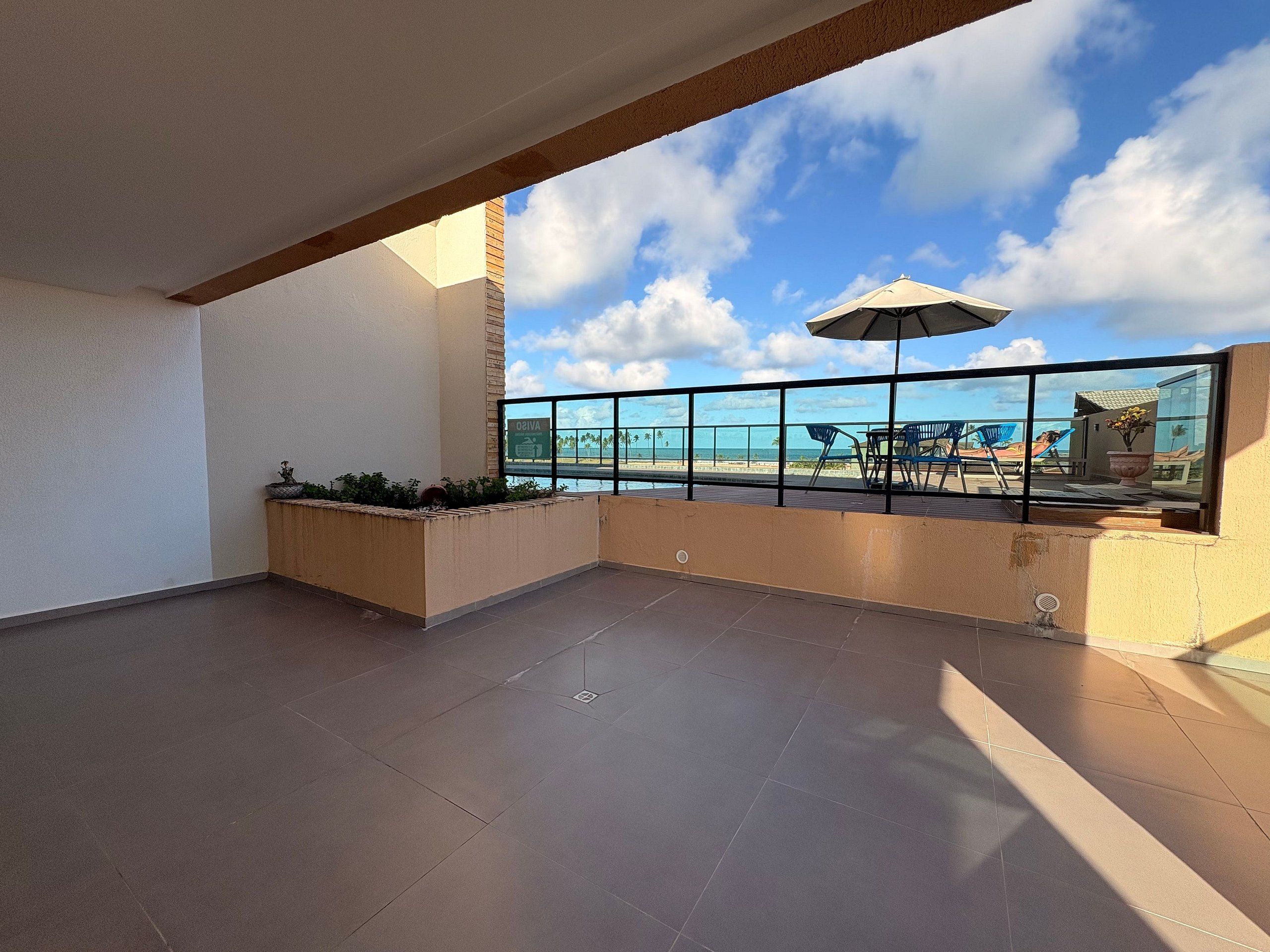 Un balcon spacieux offrant une vue dégagée sur la plage et l’océan, agrémenté d’une balustrade en verre, de mobilier d’extérieur et d’une plante en pot.