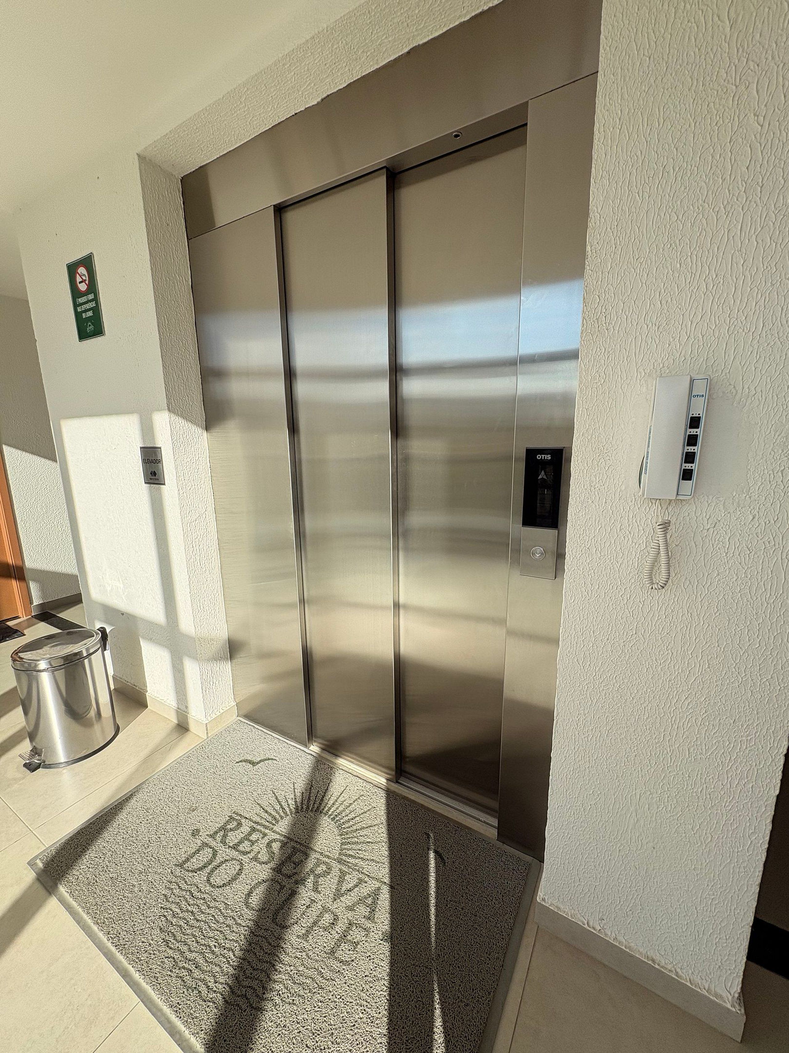 Elevador moderno com acabamento metálico, parede texturizada e uma pequena lixeira à esquerda.