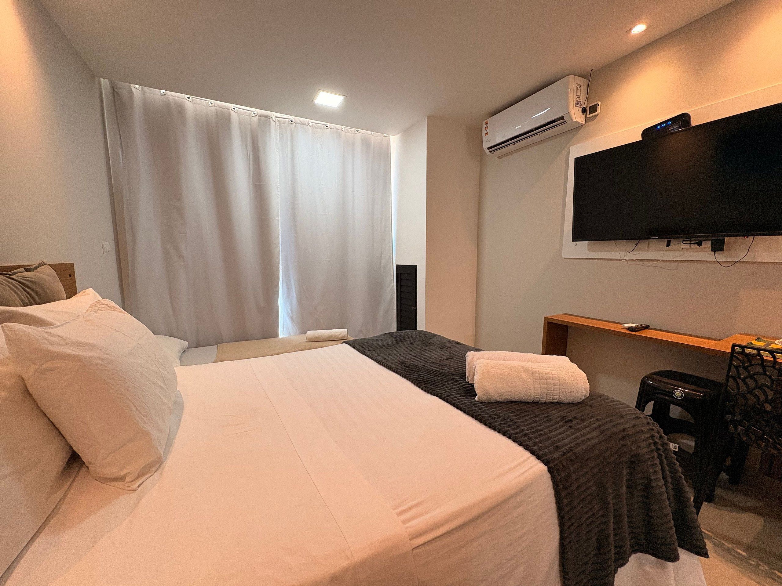 A hotel room with a bed, a TV, an air conditioning unit, and a desk.