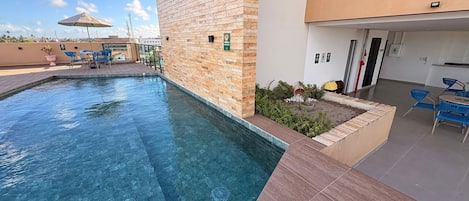 A swimming pool with a brick wall and tiled deck, surrounded by outdoor seating and a covered area.