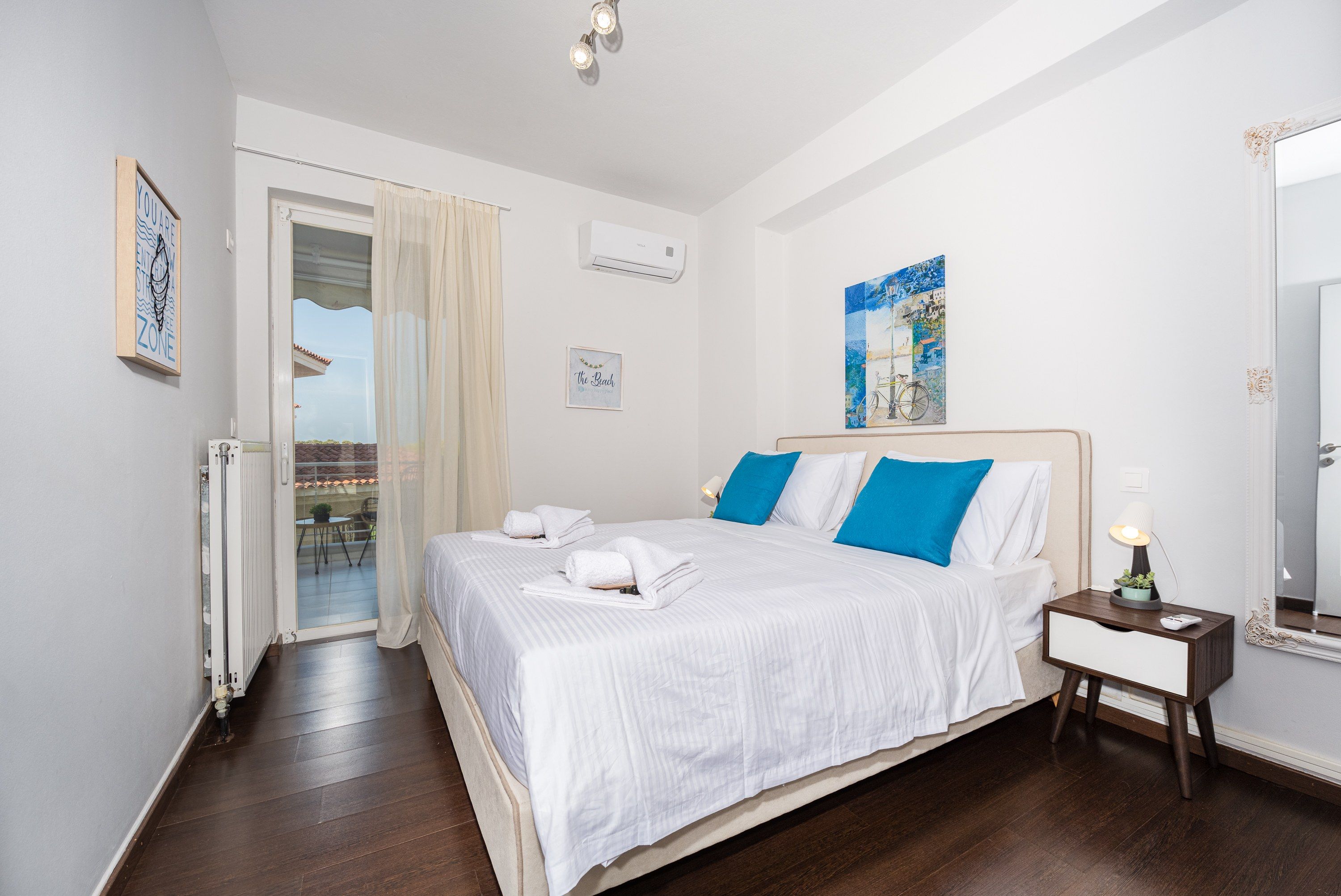 A hotel room with a bed, a nightstand, a mirror, a window with curtains, and a wall with a painting.