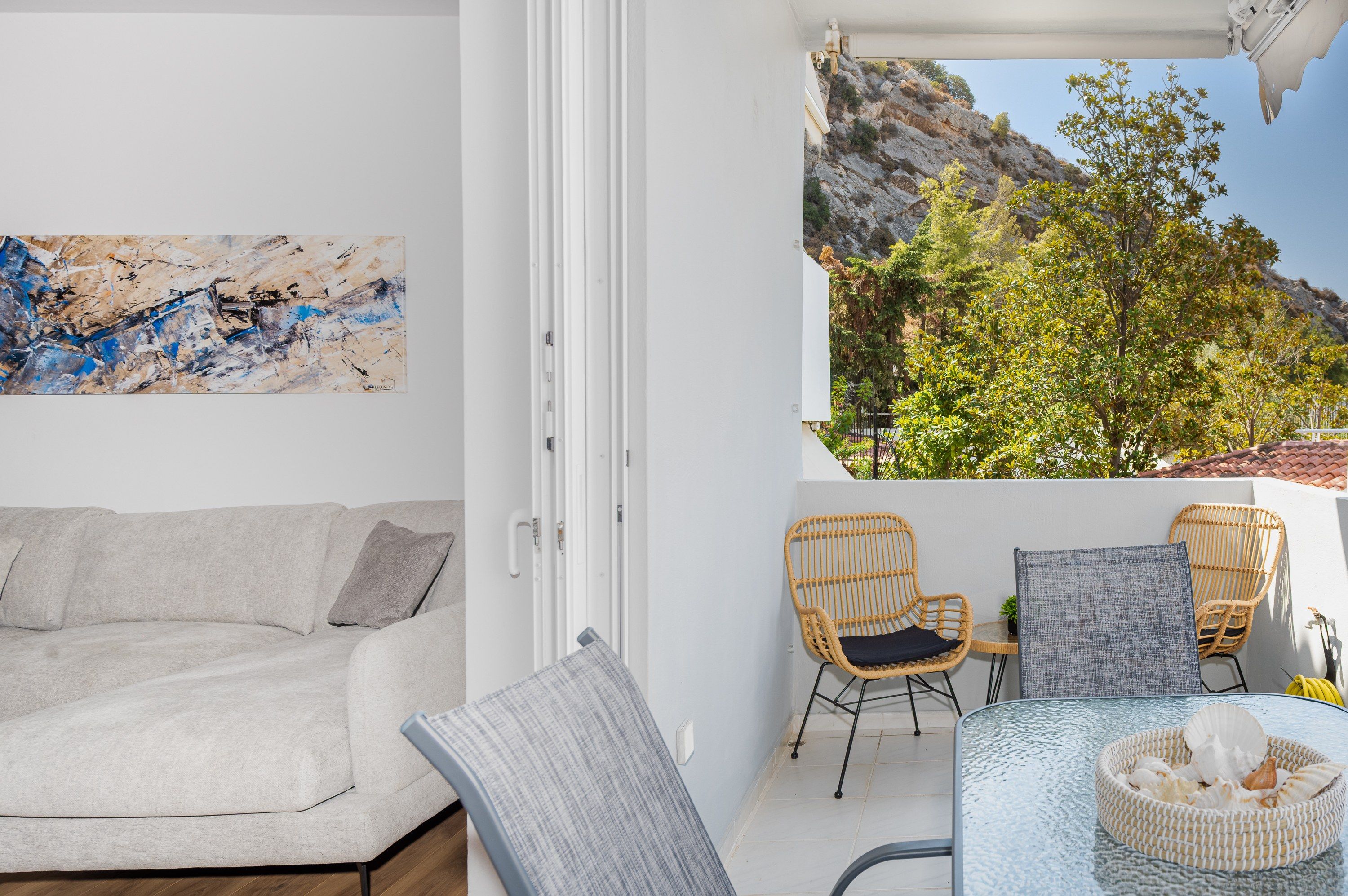 Una habitación con un sofá blanco, dos sillones de mimbre, una mesita con un cesto de conchas marinas y vistas a un paisaje rocoso con árboles.
