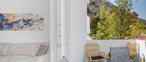 Una habitación con un sofá blanco, dos sillones de mimbre, una mesita con un cesto de conchas marinas y vistas a un paisaje rocoso con árboles.