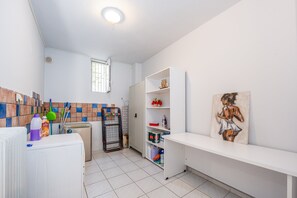 A clean, well-lit bathroom with a tiled floor, a window, a radiator, a shelf with toiletries, and a painting on the wall.