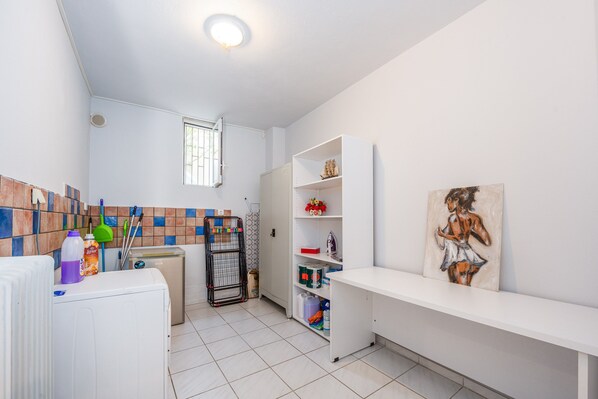 A clean, well-lit bathroom with a tiled floor, a window, a radiator, a shelf with toiletries, and a painting on the wall.