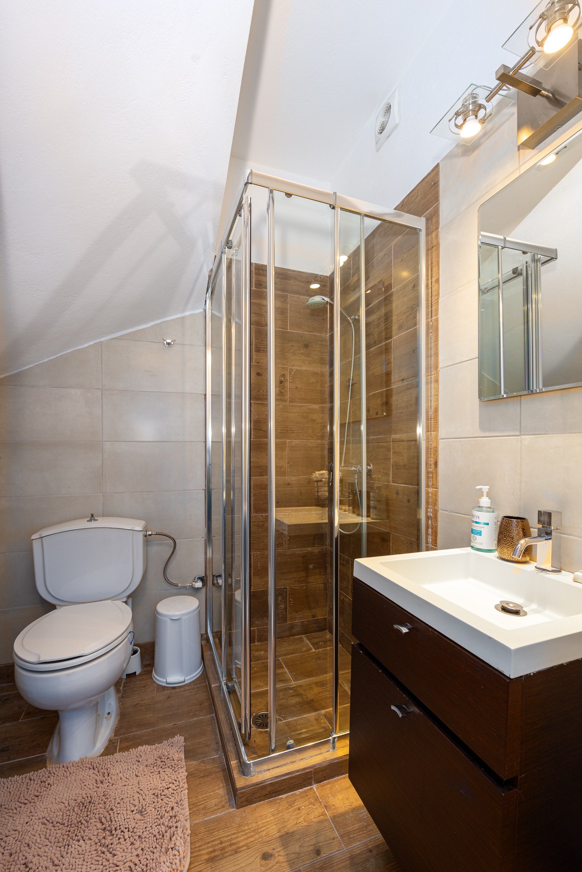 Un baño moderno con ducha envuelta en vidrio, inodoro blanco, tocador de madera y un espejo grande.