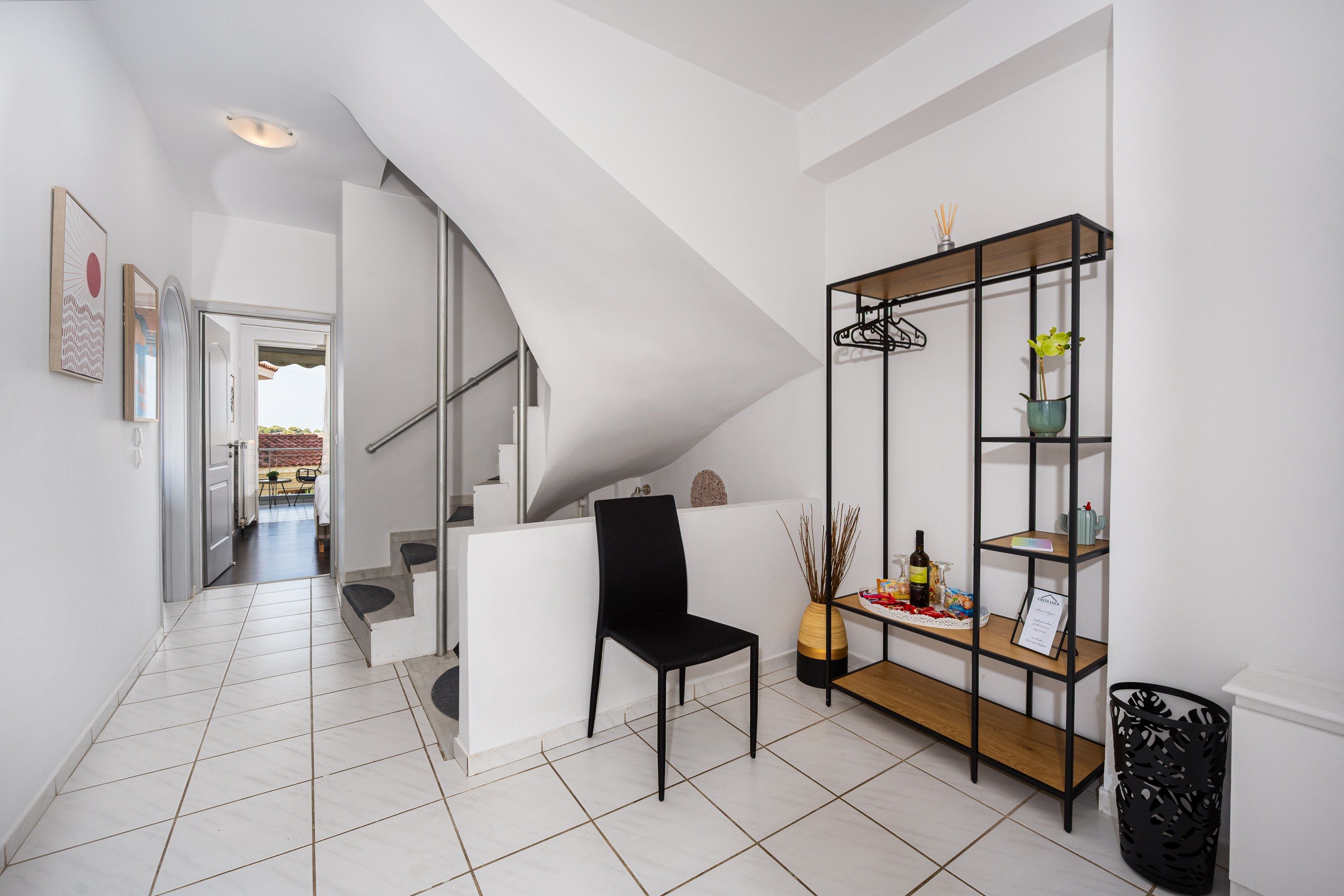 Un interior moderno con una escalera, una silla negra, un estante de madera con objetos y un suelo de baldosas.