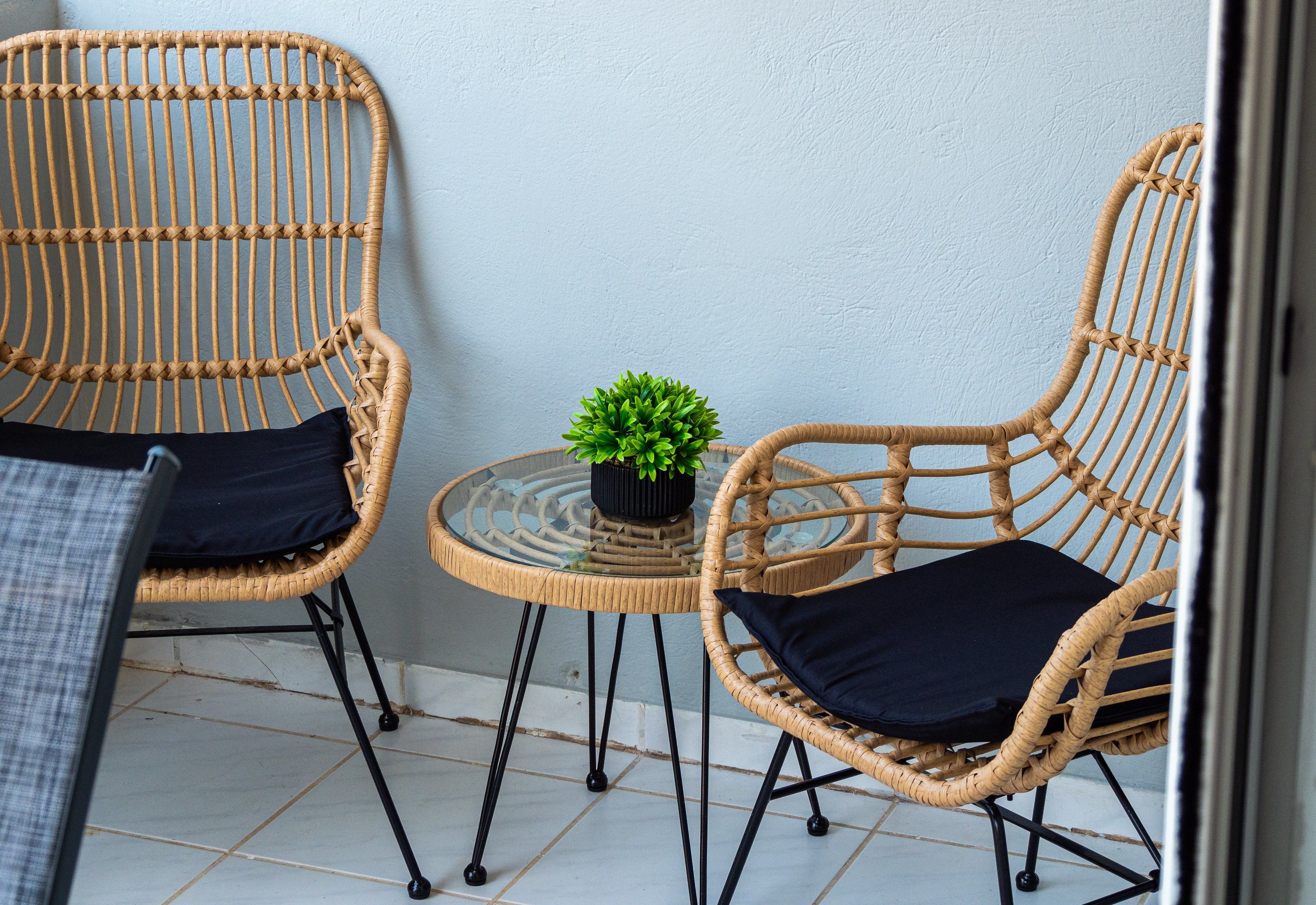Twee rieten stoelen met zwarte kussens, een kleine ronde tafel met een plant in pot, en een betegelde vloer.