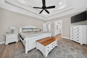 Un dormitorio con cama, mesitas de noche, un banco de madera, ventilador de techo, cómoda y televisor en la pared.