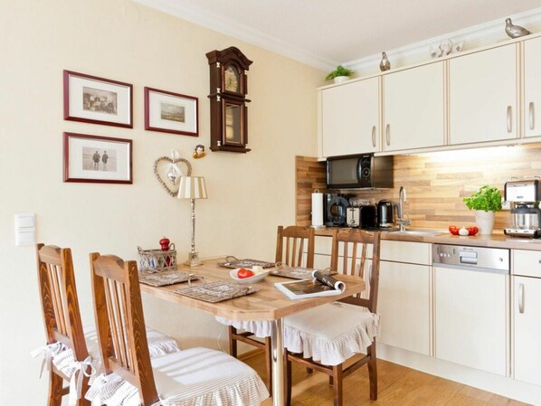 A dining area with a wooden table and chairs, a clock on the wall, framed pictures, and a kitchen with cabinets and appliances. - Holiday Apartment in Norderney With Wi-fi (Norderney)