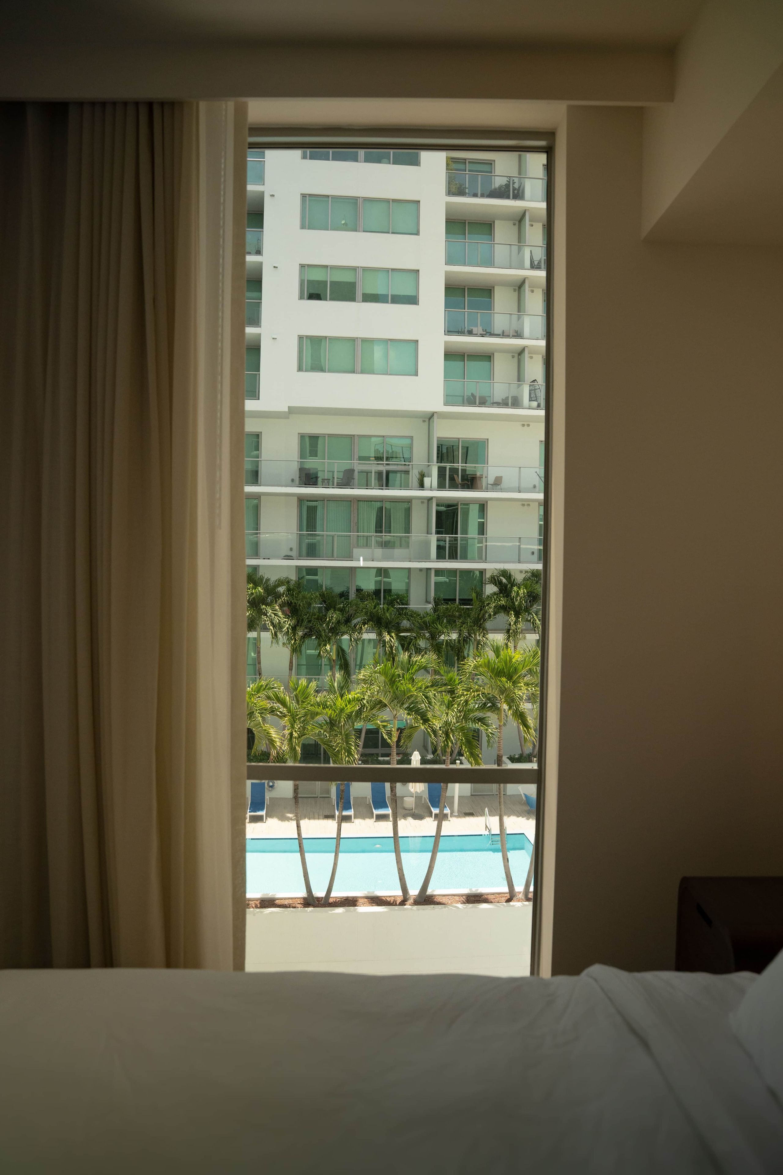 Quarto de hotel com uma janela ampla, uma cama e vista para um edifício e uma piscina.
