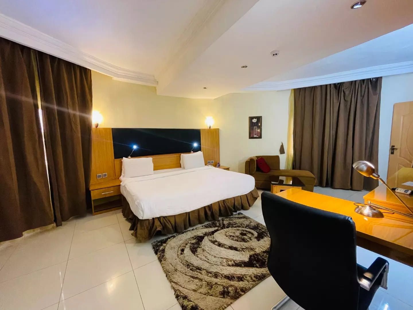 Habitación de hotel con cama, escritorio, silla, lámpara de escritorio, espejo y ventana con cortinas.