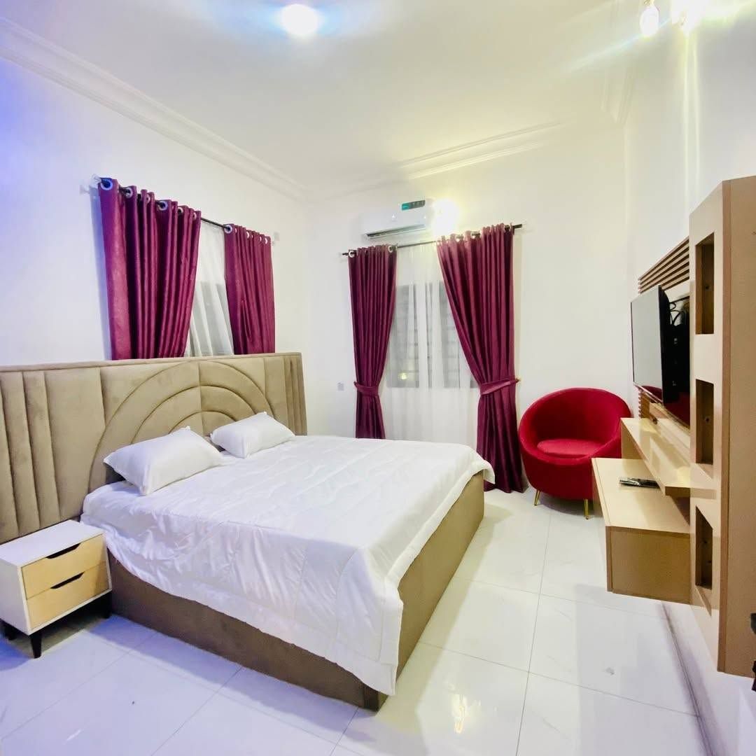 Quarto de hotel com cama, mesa de cabeceira, uma cadeira vermelha, televisão e janela com cortinas.