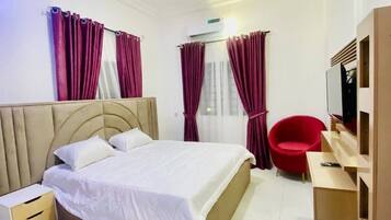 Quarto de hotel com cama, mesa de cabeceira, uma cadeira vermelha, televisão e janela com cortinas.