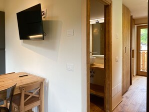 Una habitación con una mesa de comedor de madera y sillas, un televisor de pantalla plana montado en la pared, una puerta de madera que da a un baño con lavabo y espejo.