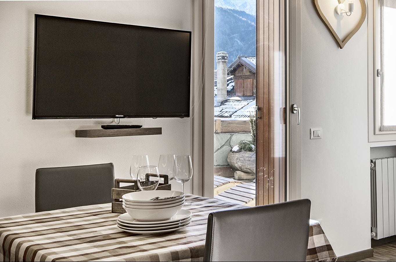 A dining area with a television mounted on the wall, a table set with plates and glasses, and a view of a mountain landscape through a glass door.