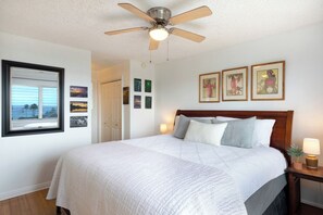 A bedroom with a bed, a ceiling fan, a nightstand with a lamp, and framed pictures on the wall. - Kailua Village#609 High Floor, Heart of Downtown, Walk to everything (Kailua Kona)