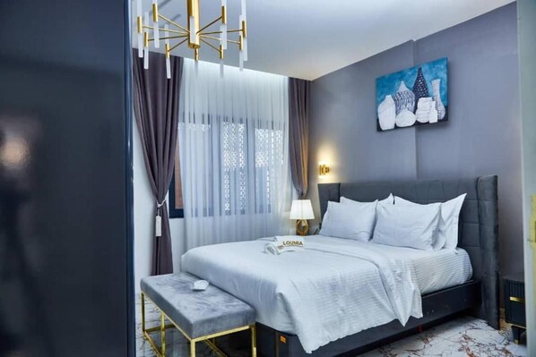 A modern hotel room with a bed, a bedside table, a bench, a painting, and a lamp.