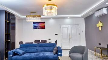 A modern living room with a blue sofa, a grey armchair, a chandelier, and a painting on the wall.