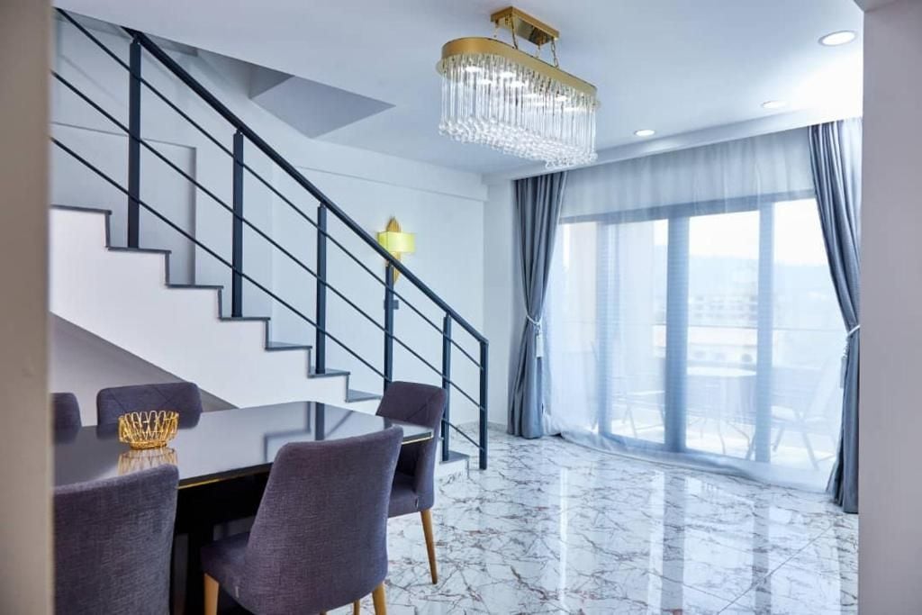 Een moderne hotelkamer met een marmeren vloer, een zithoek met een glazen tafel en stoelen, een trap met een zwarte balustrade, en een groot raam met gordijnen.