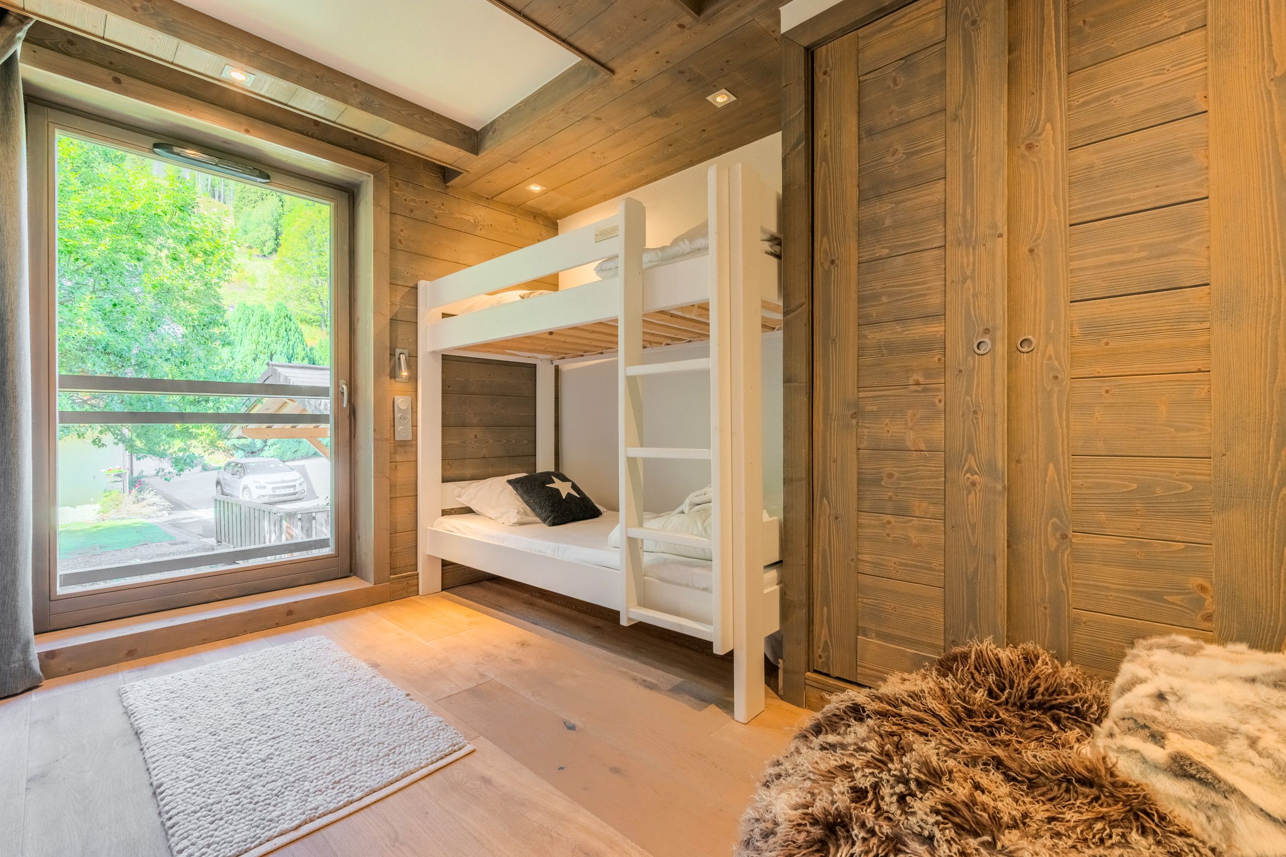 Una habitación con techo de madera, un ventanal con vistas a zonas verdes, una cama blanca con almohada y una alfombra mullida en el suelo.