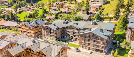 A cluster of wooden buildings with multiple balconies, surrounded by lush greenery and a hilly landscape.