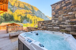 Un jacuzzi en una terraza de madera con un muro de piedra y vistas a la montaña al fondo.