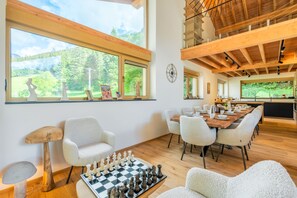 Uma sala de estar ampla com um jogo de damas, uma mesa de jantar com cadeiras e janelas grandes que oferecem vista para o verde.