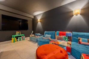 Ein modernes Wohnzimmer mit einem Ecksofa, bunten Kissen, einem grünen Spieltisch mit Spielzeug und zwei Wandleuchten.