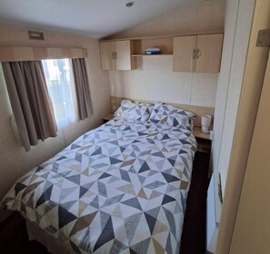 A single bed with a geometric patterned bedspread, a bedside table with a cup, and a window with curtains. - Golden Sands Haven, Sunflower Haven (Mablethorpe)