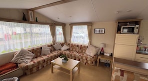 A cozy living room with a sectional sofa, a small table, and a kitchenette area with a microwave. - Golden Sands Haven, Sunflower Haven (Mablethorpe)