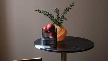 Una mesa redonda con un florero que contiene manzanas rojas y follaje verde, un recipiente negro y una silla de madera con un cojín azul.