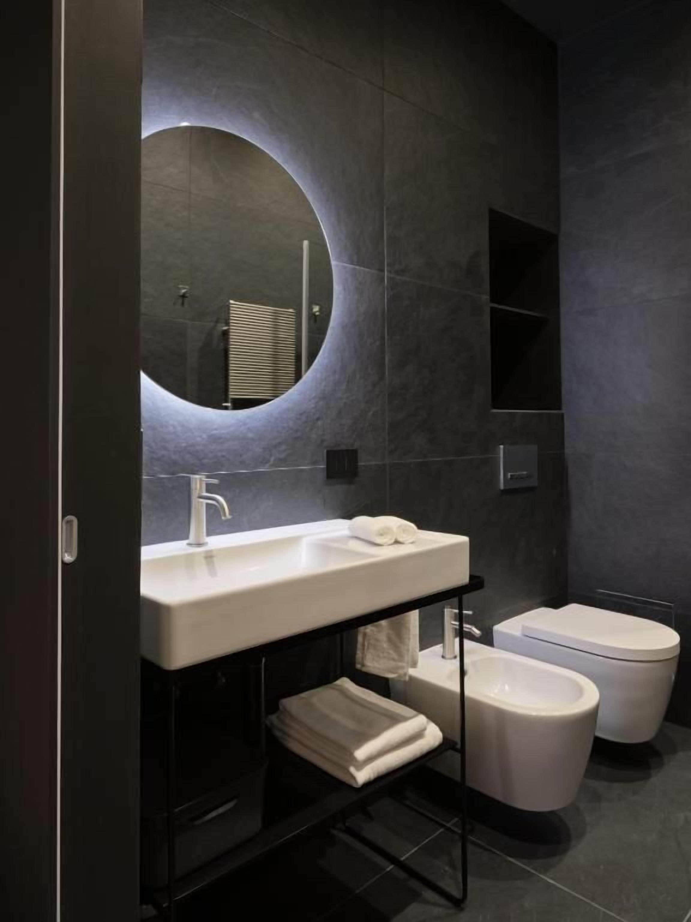 Une salle de bain moderne avec un grand miroir rond, un lavabo blanc, des toilettes et un bidet.