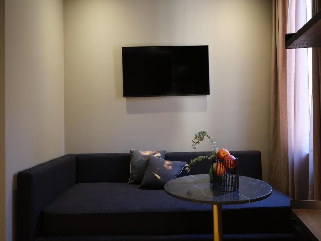 Una sala de estar moderna con un sofá oscuro, un televisor de pantalla plana montado en la pared y una pequeña mesa redonda con un jarrón de flores.