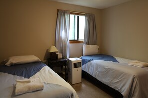 A hotel room with two single beds, a bedside table with a lamp, a window with curtains, and an air conditioning unit. - Private home in Waterville Estates! (Thornton)