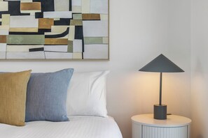 Una cama con dos almohadas, una lámpara moderna sobre una mesita de noche y un cuadro abstracto geométrico en la pared.