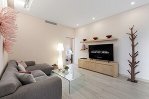 A modern living room with a flat-screen TV, grey sofa, glass coffee table, and a wooden coat rack. - 3BR Playacar 3rd Floor - Rooftop Pool, Gym, Gated 24/7 (Playa del Carmen)