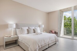A modern bedroom with a large bed, bedside tables, a nightstand, a chair, and a large window offering a view of greenery. - 3BR Playacar 3rd Floor - Rooftop Pool, Gym, Gated 24/7 (Playa del Carmen)